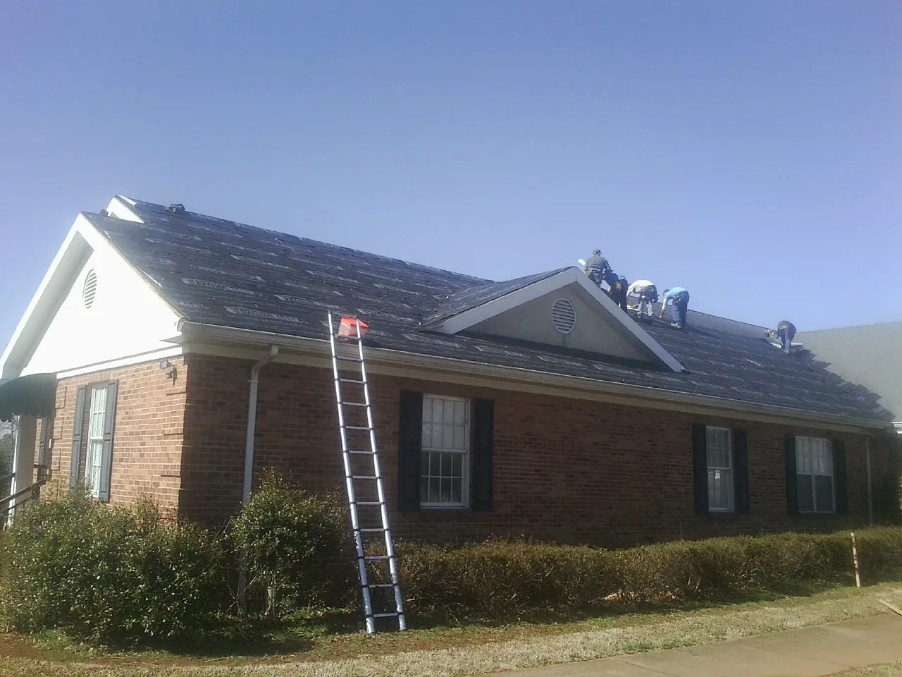Crew tearing shingles off a brick ranch roof with a ladder set — Metal Roof Repair work by our crew in Brookville